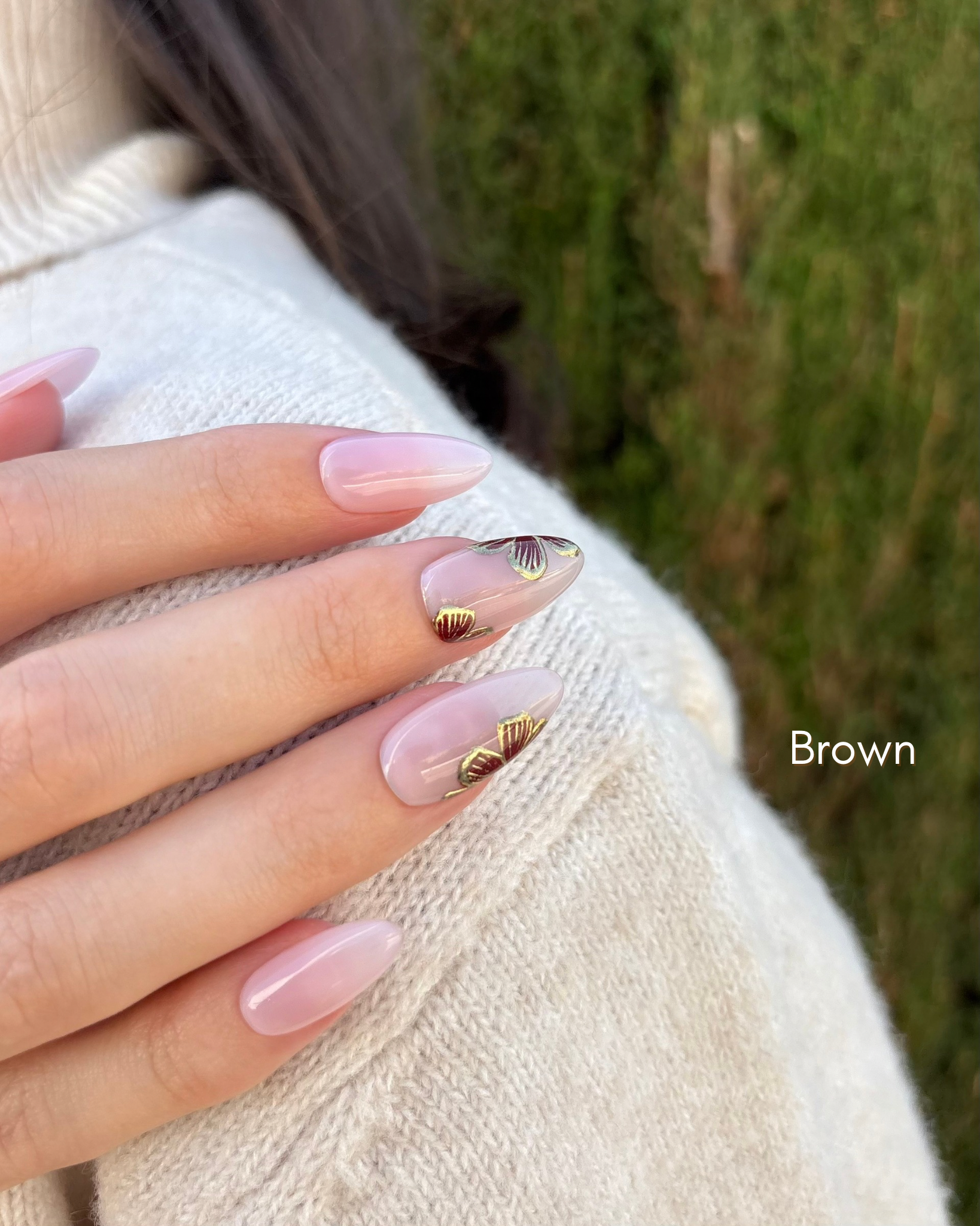 mano con uñas press-on puestas nudes con flores en color marrón y contorno dorado cromático apoyada sobre el hombro