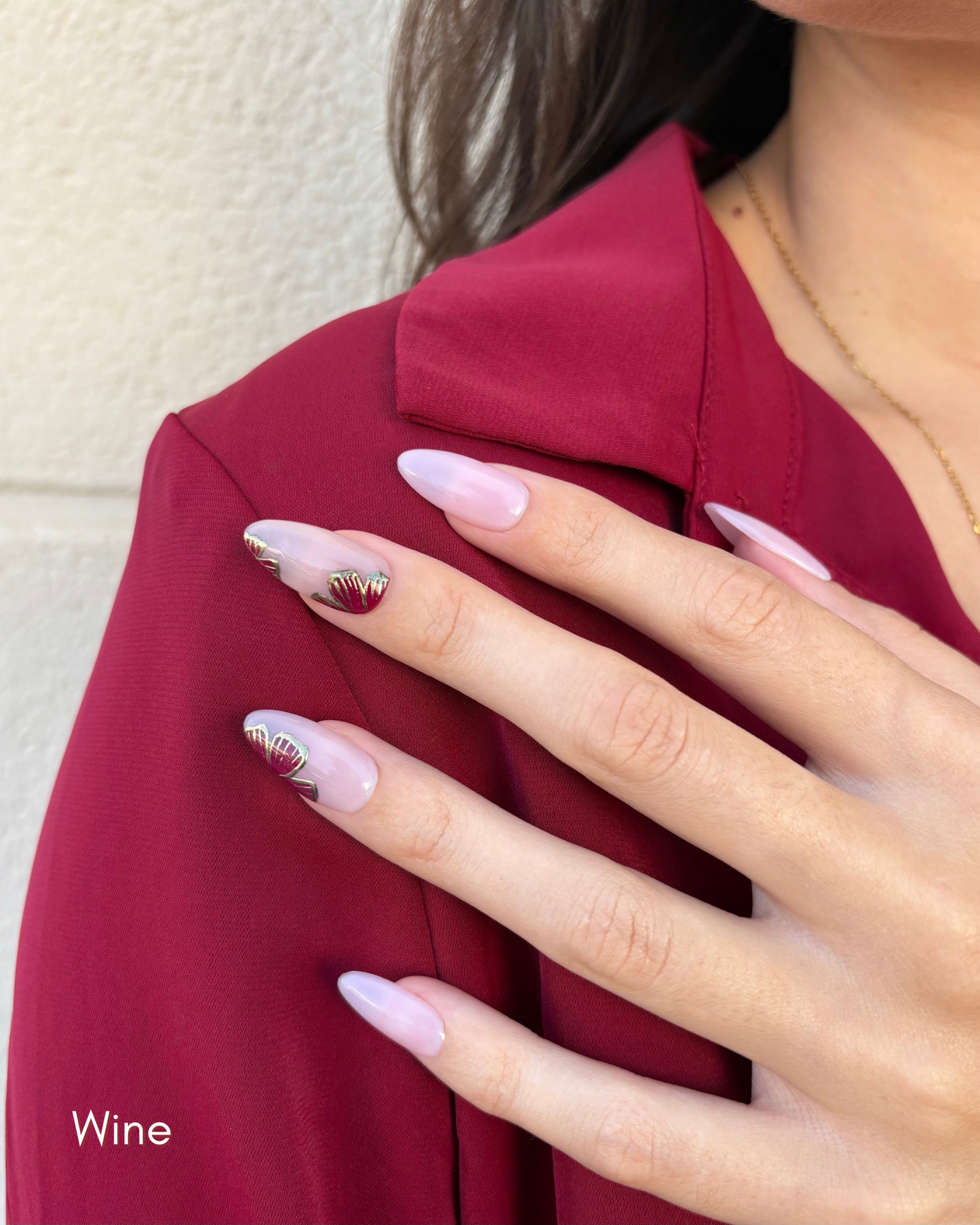una mano con uñas press-on nudes con flores  en color vino y contorno dorado cromático y camisa burdeos