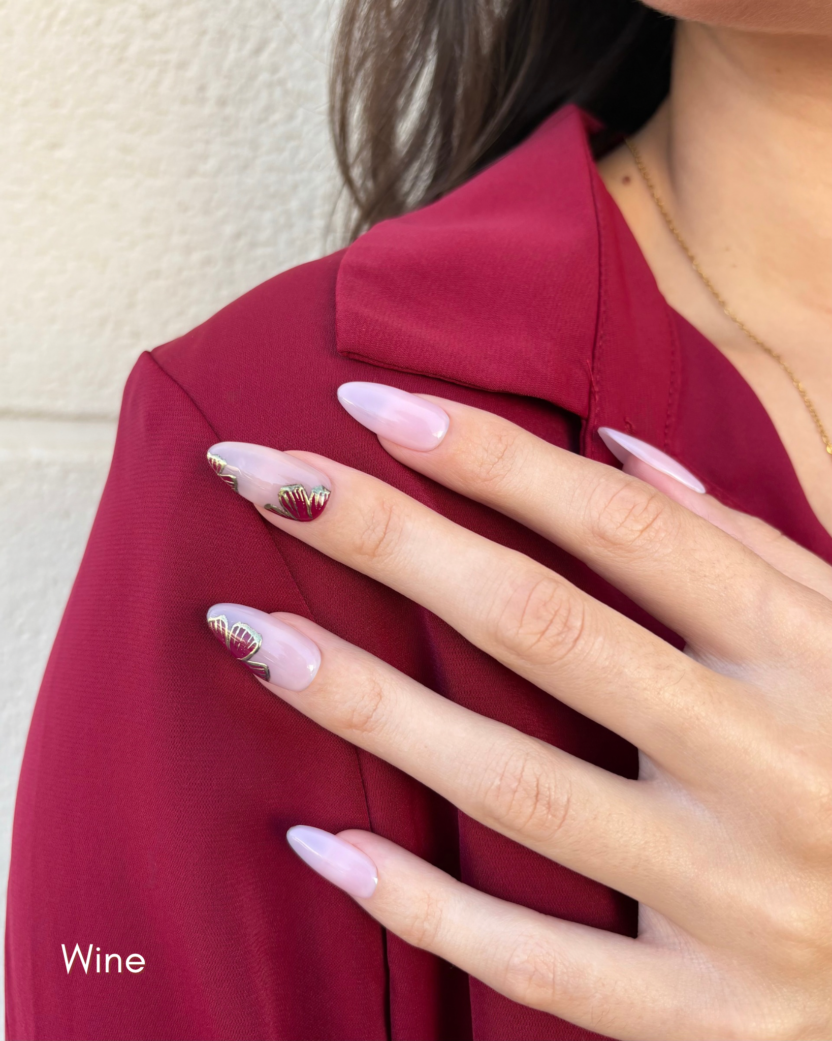 una mano con uñas press-on nudes con flores  en color vino y contorno dorado cromático y camisa burdeos