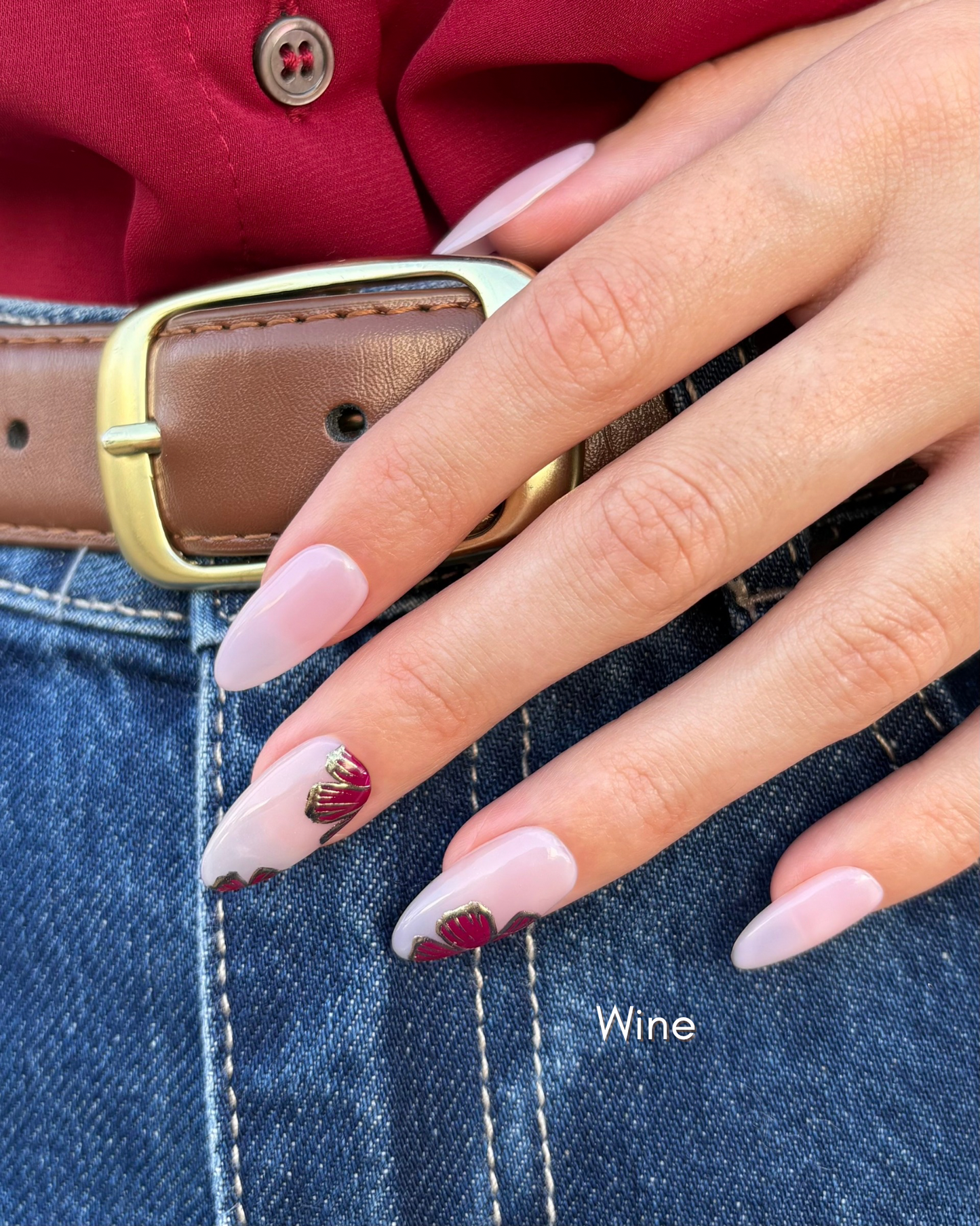 mano con uñas press-on puestas nudes con flores en color vino y contorno dorado cromático apoyada en el pantalón con cinturón