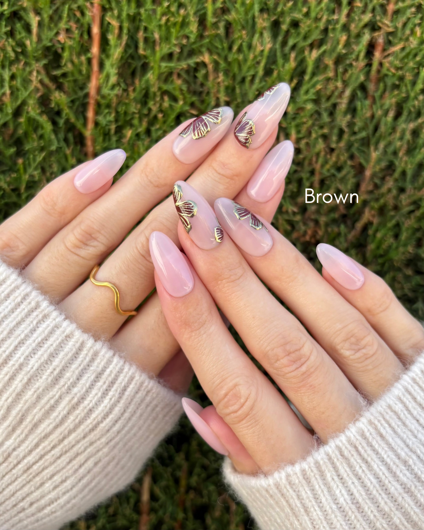 mano con uñas press-on puestas nudes con flores en color marrón y contorno dorado cromático