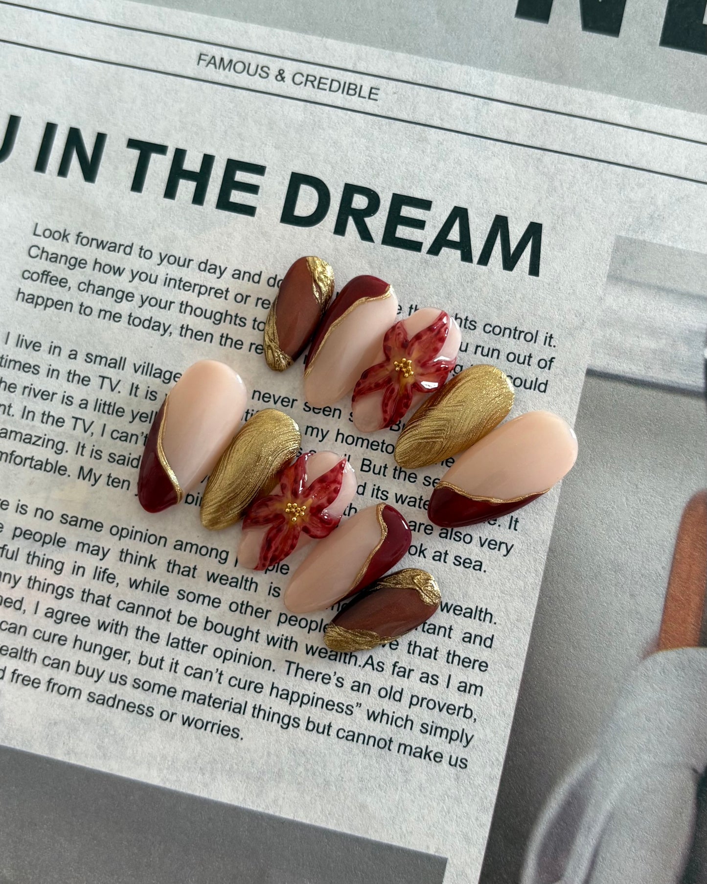 set de uñas press-on otoñal dorado y rojo burdeos con ojo de gato, cromático y flor 3D puestas sobre un periodico