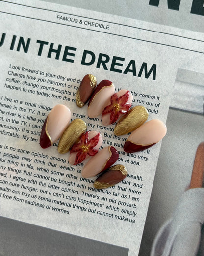set de uñas press-on otoñal dorado y rojo burdeos con ojo de gato, cromático y flor 3D puestas sobre un periodico