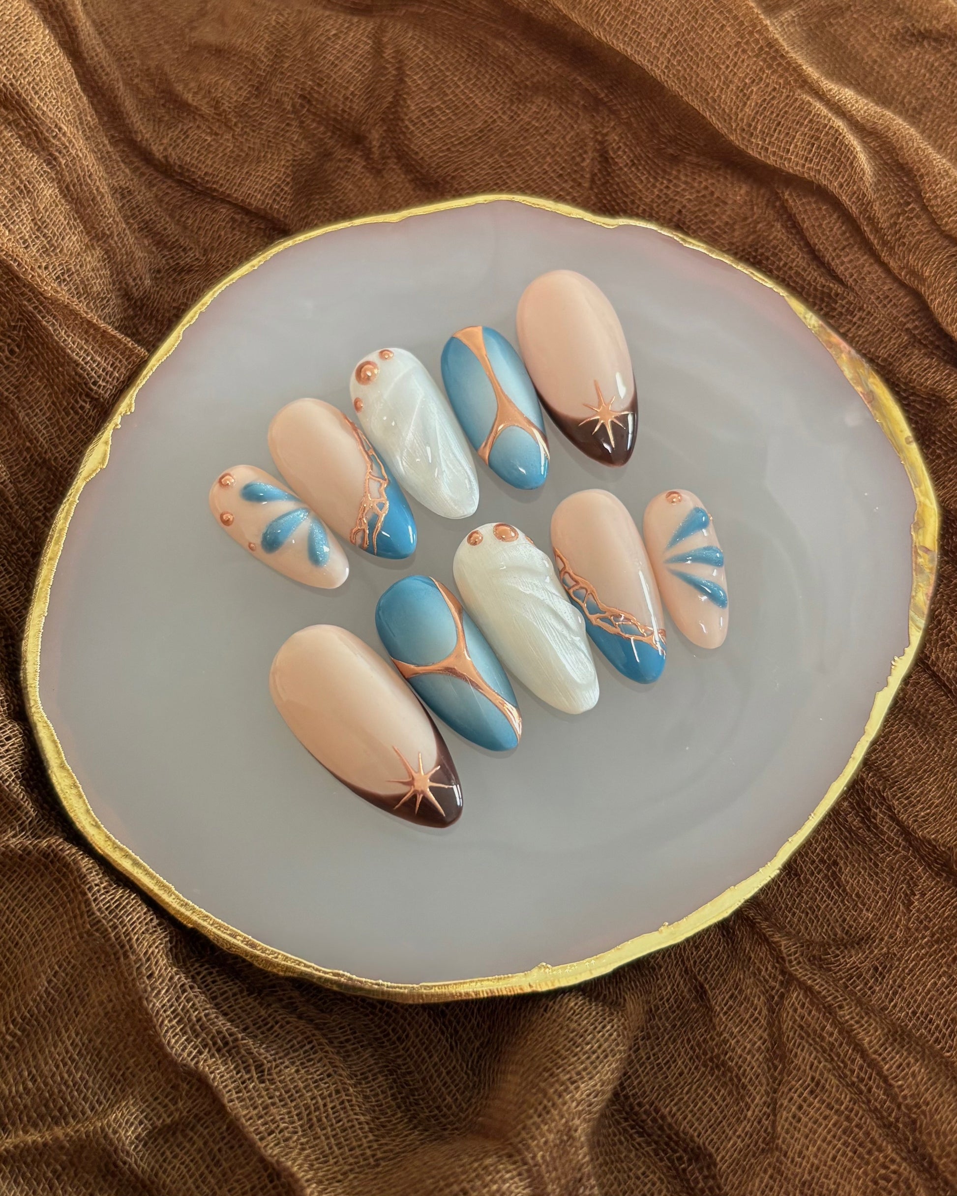 set de uñas press-on azul con toques de cobre cromático, degradado con aerógrafo, blanco perlado con gotas de gel 3D, ojo de gato y estrella sobre paleta de uñas blanca y mantel marrón