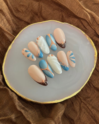 set de uñas press-on azul con toques de cobre cromático, degradado con aerógrafo, blanco perlado con gotas de gel 3D, ojo de gato y estrella sobre paleta de uñas blanca y mantel marrón