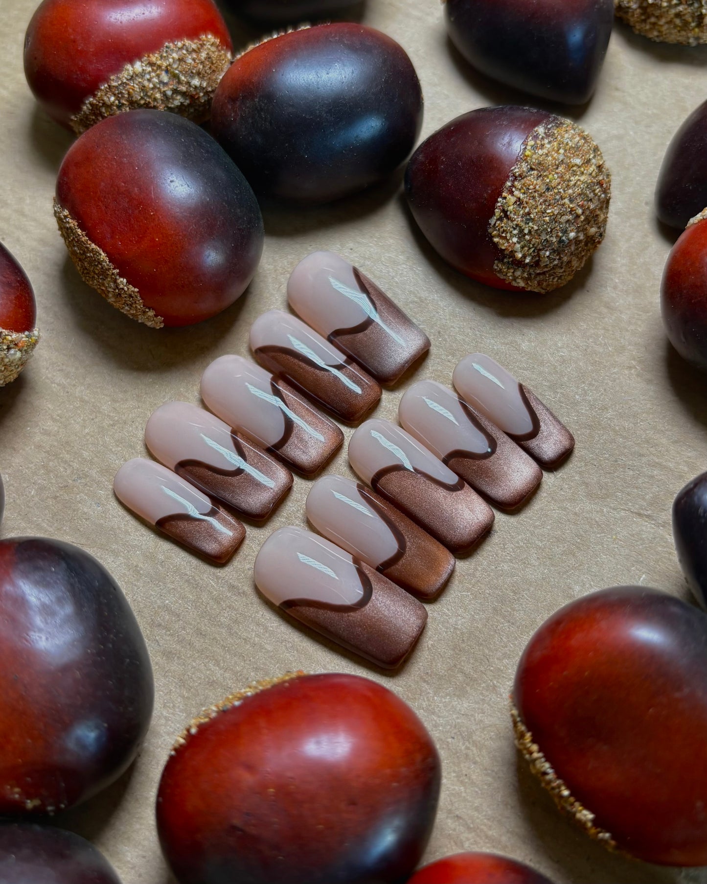 set de uñas press-on nude con francesa abstracta de color marrón con efecto ojo de gato y un línea delgada marrón que separa la francesa con castañas alrededor