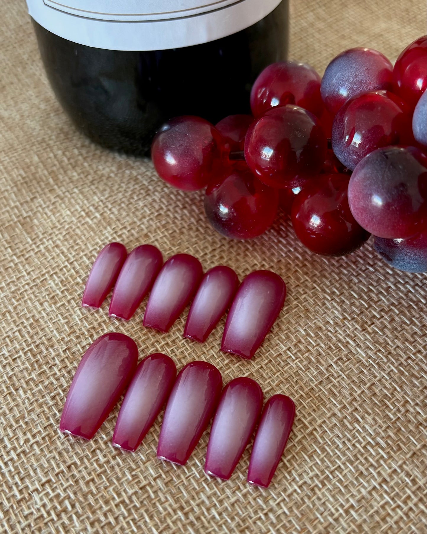 set de uñas press-on burdeos con degradado aura en el centro de color beige hecho con aerógrafo puestas sobre una tela beige , al lado de uva y una botella de vino