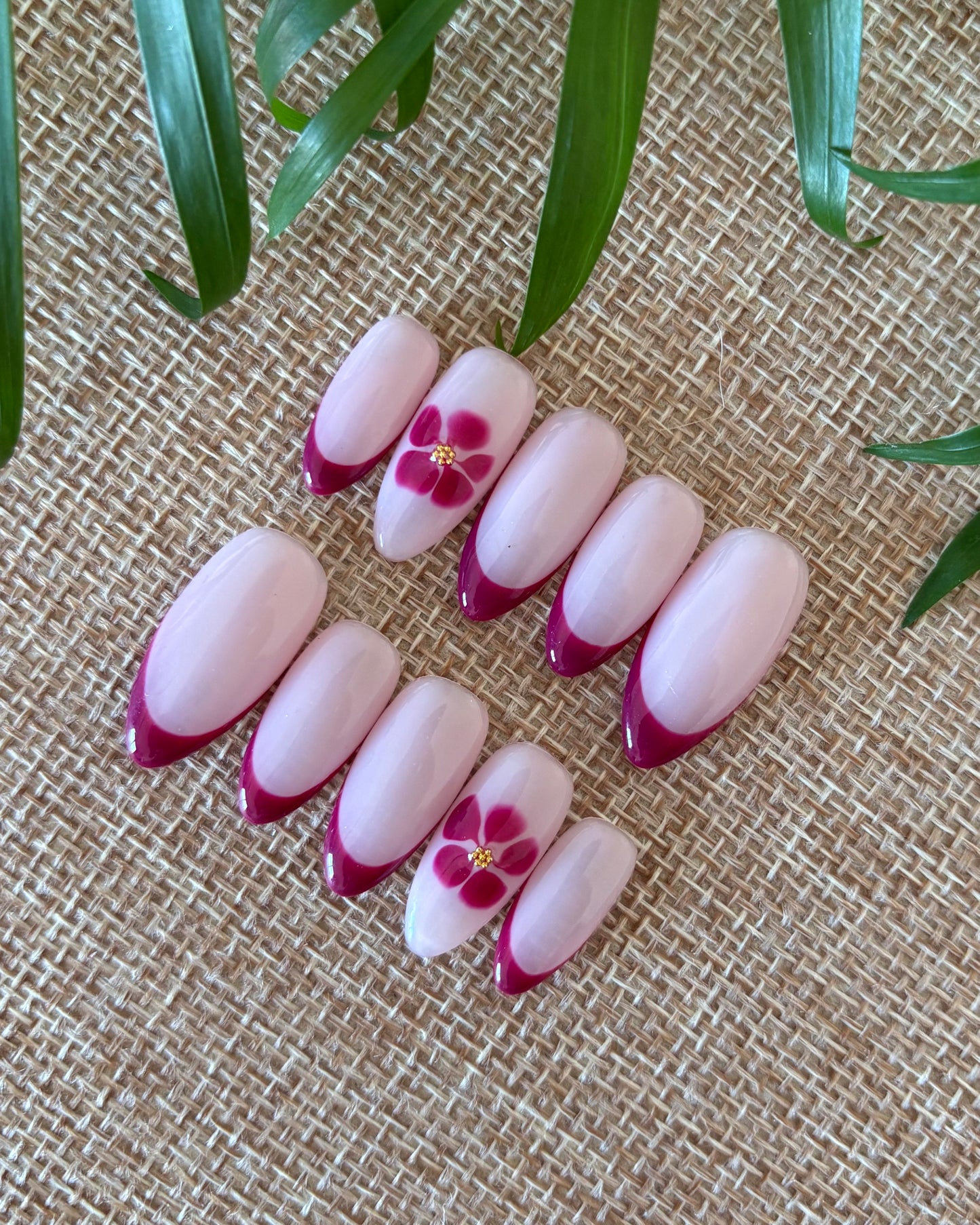 set de uñas press-on nudes con francesa burdeos y flor blooming con caviar dorado sobre fondo de tela beige y planta