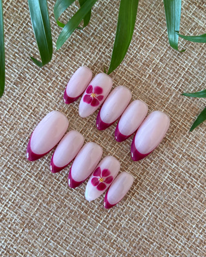 set de uñas press-on nudes con francesa burdeos y flor blooming con caviar dorado sobre fondo de tela beige y planta