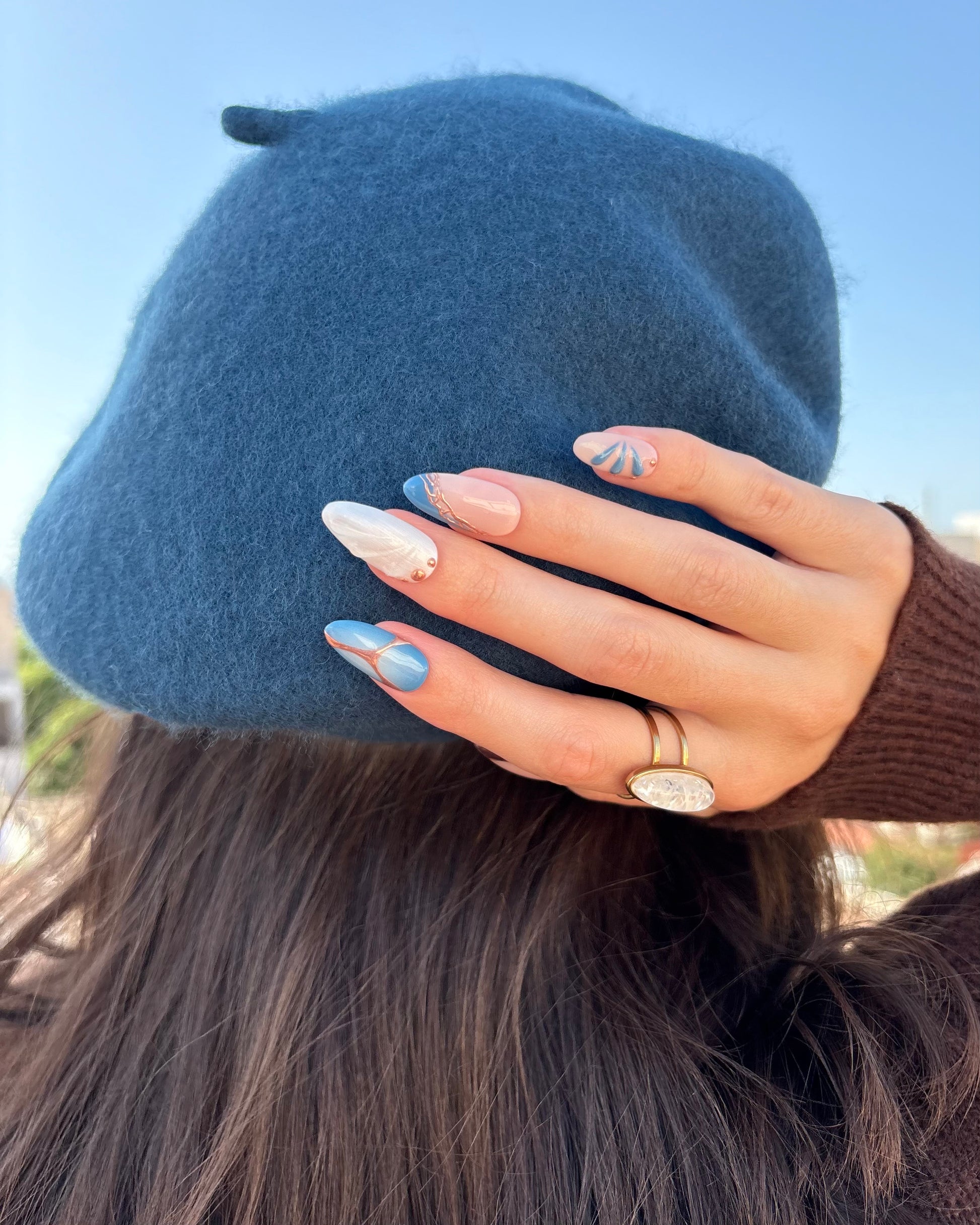 set de uñas press-on azul con toques de cobre cromático, degradado con aerógrafo, blanco perlado con gotas de gel 3D, ojo de gato y estrella una mano apoyada sobre beret azul pato