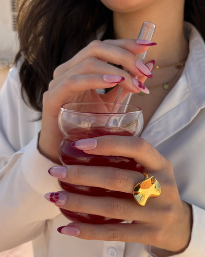 set de uñas press-on nudes con francesa burdeos y flor blooming con caviar dorado cogiendo un bubble tea con anillos