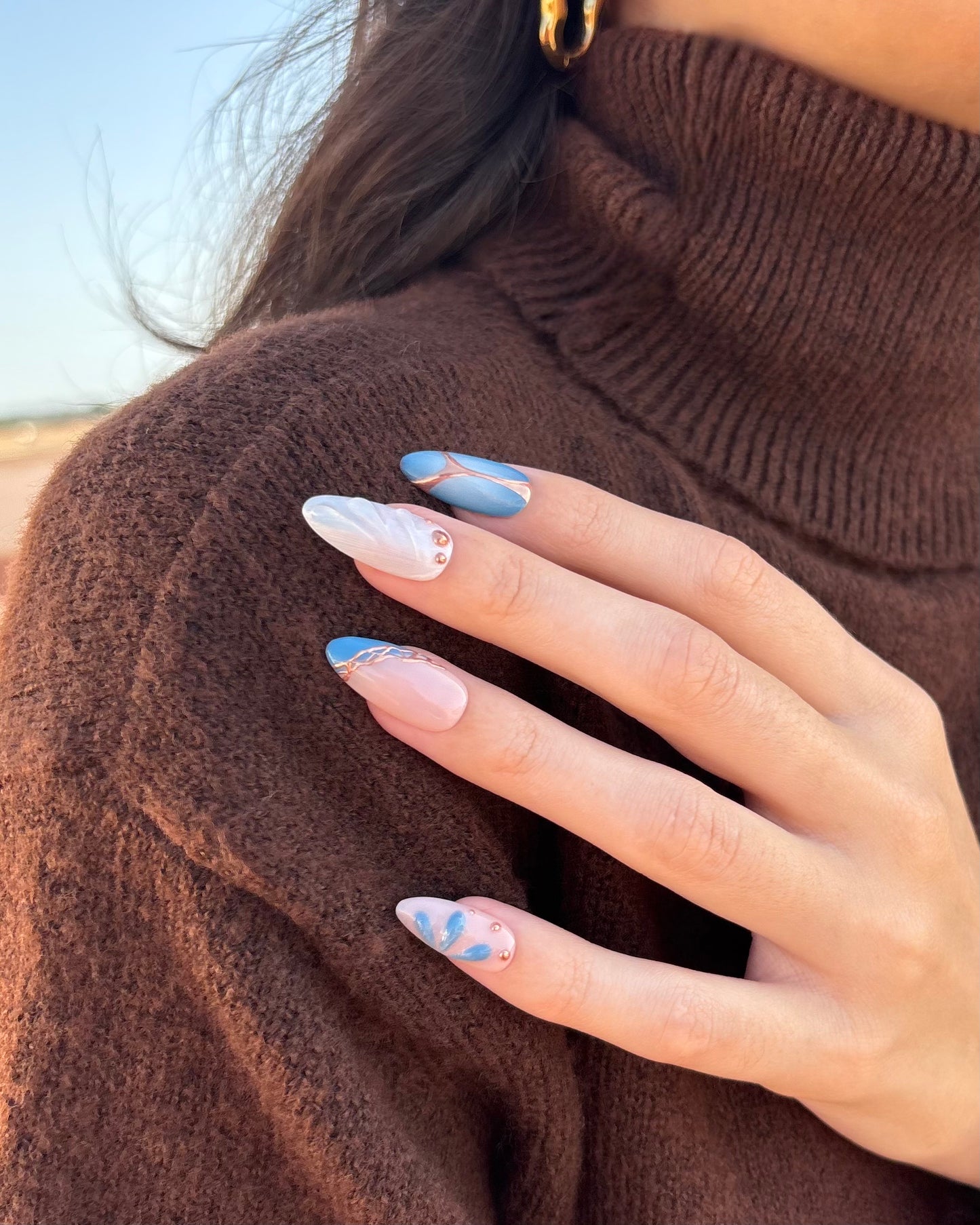 set de uñas press-on azul con toques de cobre cromático, degradado con aerógrafo, blanco perlado con gotas de gel 3D, ojo de gato y estrella apoyadas en hombro con jersey marrón