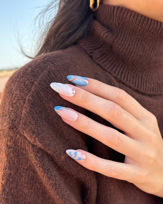 set de uñas press-on azul con toques de cobre cromático, degradado con aerógrafo, blanco perlado con gotas de gel 3D, ojo de gato y estrella apoyadas en hombro con jersey marrón