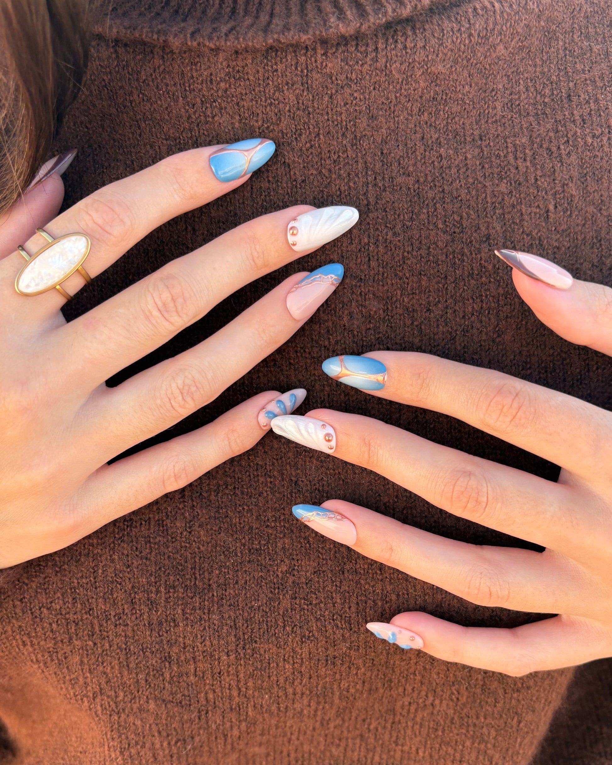 set de uñas press-on azul con toques de cobre cromático, degradado con aerógrafo, blanco perlado con gotas de gel 3D, ojo de gato y estrella 2 manos sobre jersey marrón