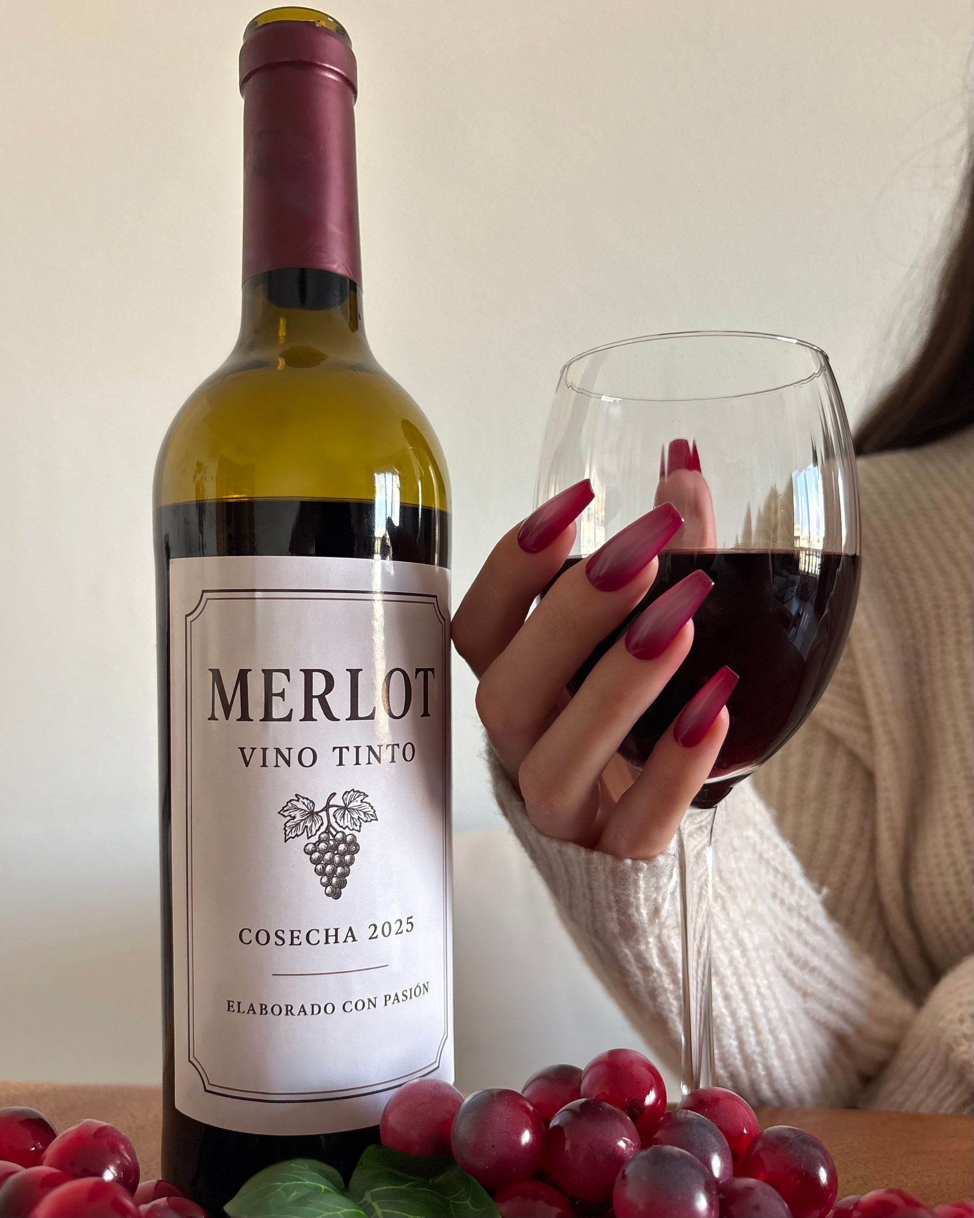 set de uñas press-on burdeos con degradado aura en el centro de color beige hecho con aerógrafo cogiendo una copa de vino con una botella de vino merlot al lado y uvas