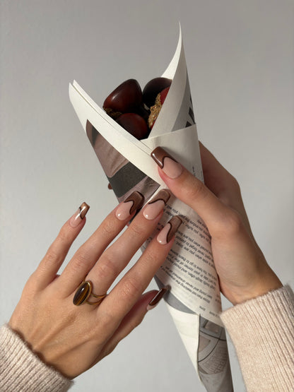 set de uñas press-on nude con francesa abstracta de color marrón con efecto ojo de gato y un línea delgada marrón que separa la francesa cogiendo un cono de castañas con fondo blanco