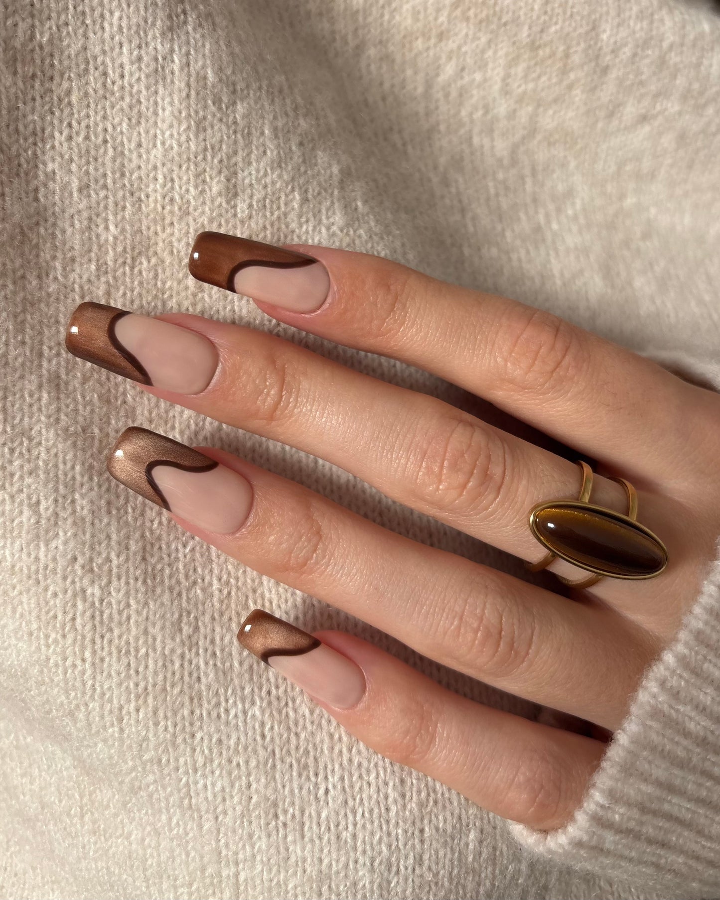 set de uñas press-on nude con francesa abstracta de color marrón con efecto ojo de gato y un línea delgada marrón que separa la francesa solo una mano con jersey beige de fondo