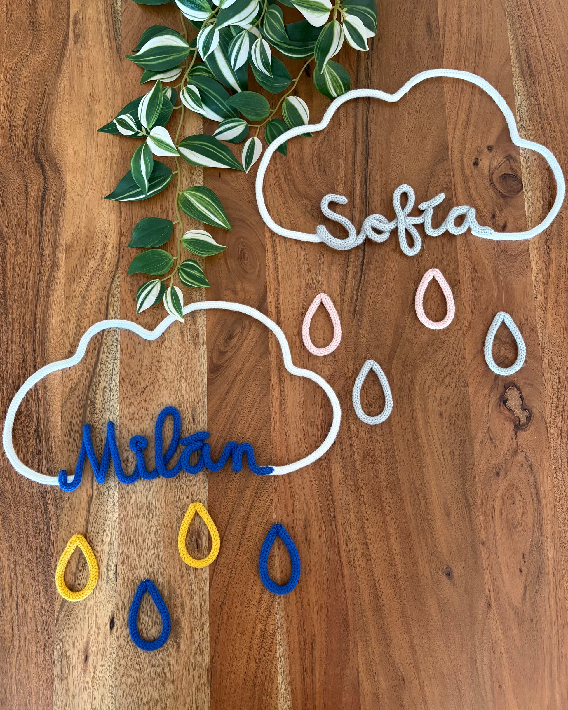 dos móviles decorativos de nube con gotas y nombres personalizados con fondo madera y planta