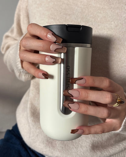 set de uñas press-on nude con francesa abstracta de color marrón con efecto ojo de gato y un línea delgada marrón que separa la francesa agarrando una taza thermo beige de café nespresso